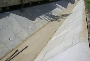 Concrete drainage channel with sloped, reinforced walls; dirt base; two metal pipes resting on the lower edge; shadows from a nearby structure or vehicle cross the scene.